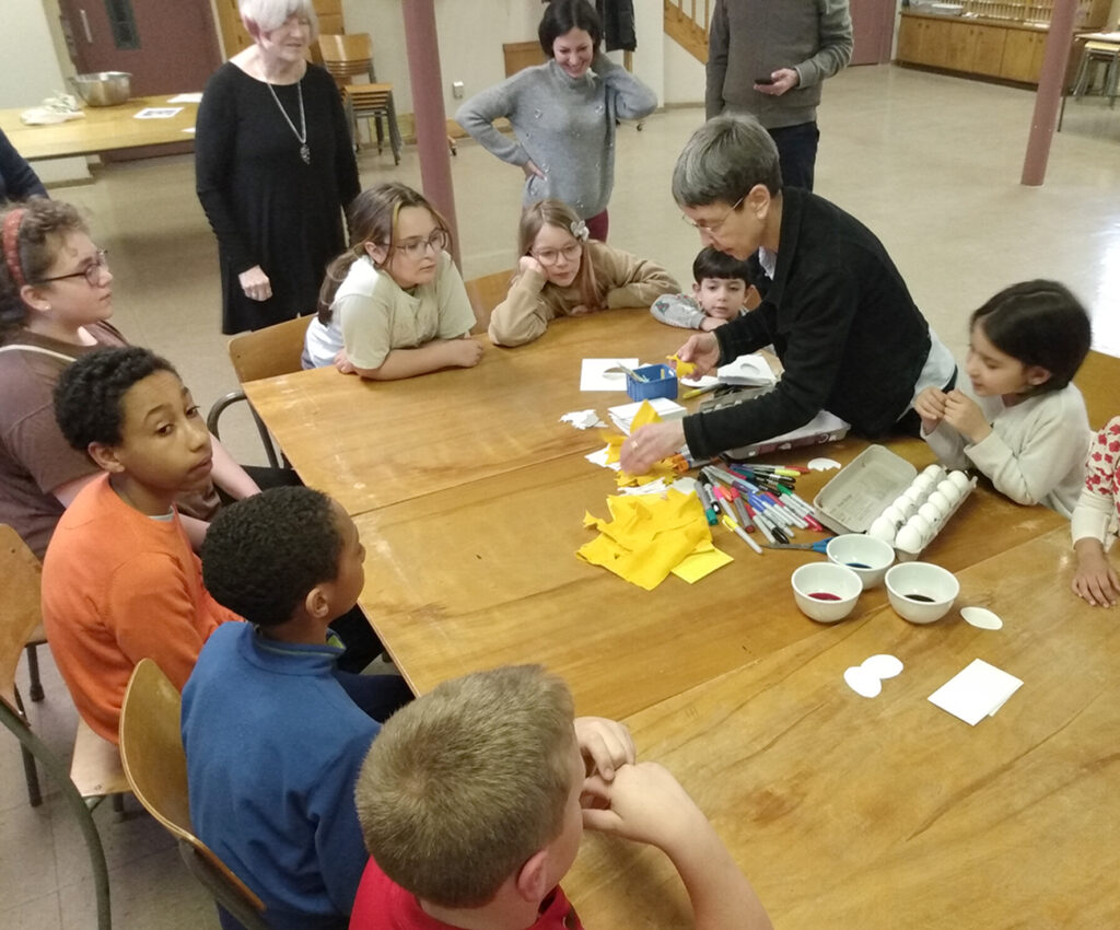 Pastor Dani is with a group of learning children that are sitting around a wooden table. There are eggs and dye and markers in the middle of the table for Easter egg decorating.
