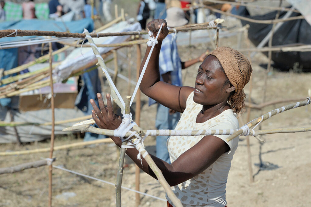 In Haiti, a woman ties binding around poles as she works on building a structure after an earthquake